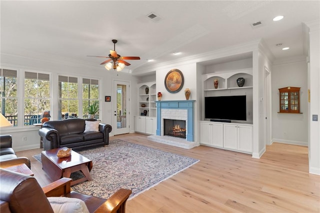 living room featuring light wood-type flooring, ornamental molding, built in features, ceiling fan, and a fireplace