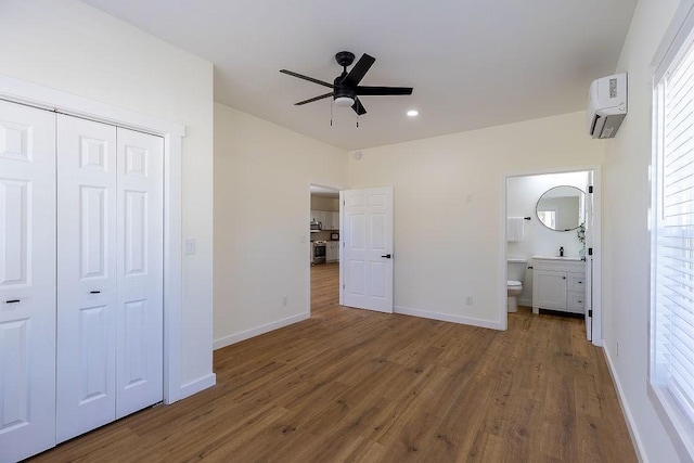 unfurnished bedroom featuring a closet, a wall unit AC, wood finished floors, ensuite bath, and baseboards
