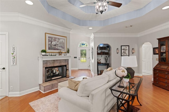 living room featuring a tiled fireplace, ceiling fan, crown molding, arched walkways, and wood finished floors
