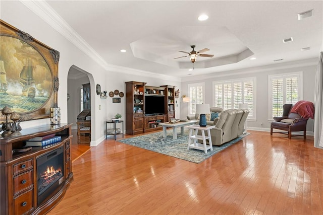 living area featuring ceiling fan, light wood-style floors, a tray ceiling, baseboards, and arched walkways