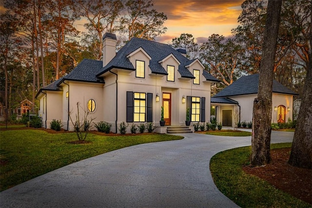 french provincial home featuring stucco siding, curved driveway, a front yard, a shingled roof, and a chimney