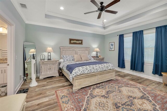 bedroom with ceiling fan, a raised ceiling, light hardwood / wood-style flooring, ensuite bathroom, and crown molding