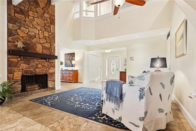 living area featuring stone tile floors, baseboards, ceiling fan, a high ceiling, and a stone fireplace