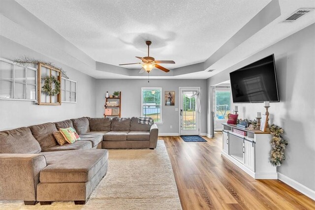 living room with a raised ceiling, ceiling fan, a textured ceiling, and light wood-type flooring