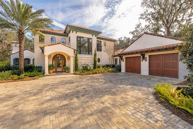 mediterranean / spanish-style home with an attached garage, a tile roof, french doors, decorative driveway, and stucco siding