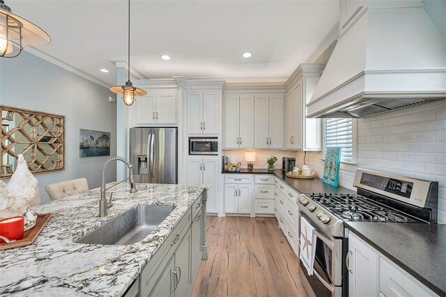 kitchen with stainless steel appliances, a sink, premium range hood, light wood-style flooring, and pendant lighting