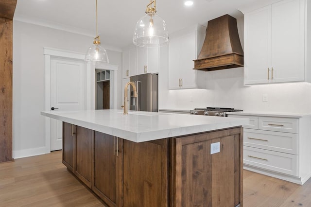 kitchen with high end fridge, custom exhaust hood, pendant lighting, white cabinetry, and an island with sink