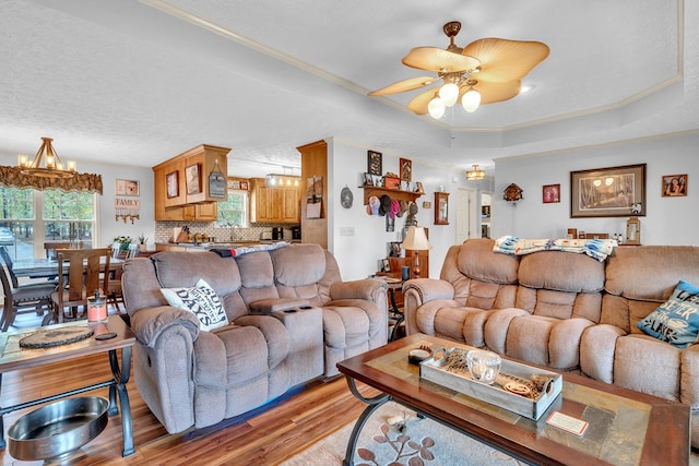 living room with a raised ceiling, light hardwood / wood-style flooring, a textured ceiling, ceiling fan with notable chandelier, and ornamental molding