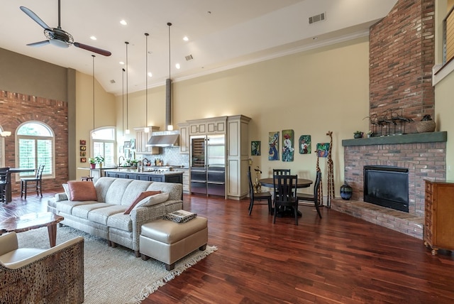 living room with a high ceiling, dark hardwood / wood-style flooring, a brick fireplace, and ceiling fan