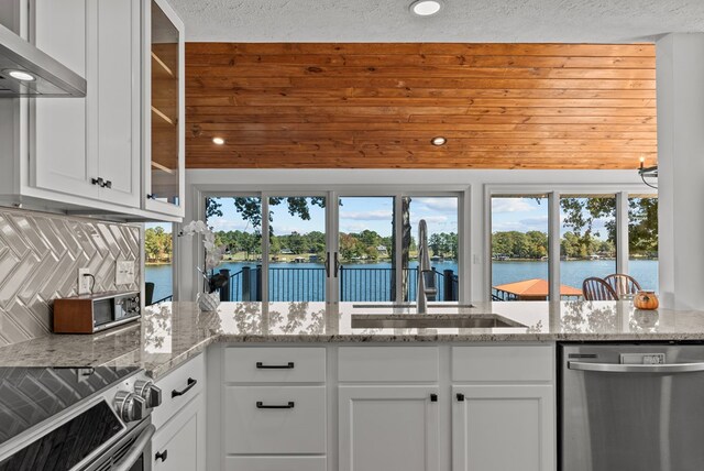 kitchen with light stone countertops, wall chimney range hood, white cabinets, a water view, and appliances with stainless steel finishes