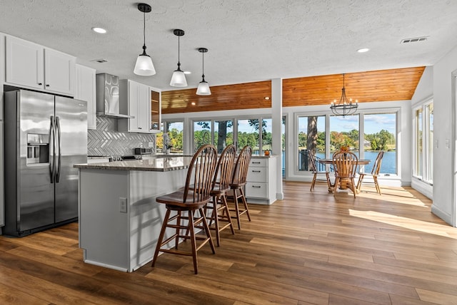 kitchen with wall chimney exhaust hood, tasteful backsplash, stainless steel fridge with ice dispenser, dark stone countertops, and white cabinets