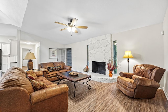 living area featuring baseboards, ceiling fan, wood finished floors, vaulted ceiling, and a stone fireplace