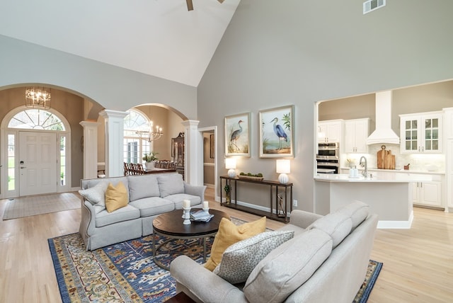 living room featuring ornate columns, sink, high vaulted ceiling, a chandelier, and light hardwood / wood-style floors