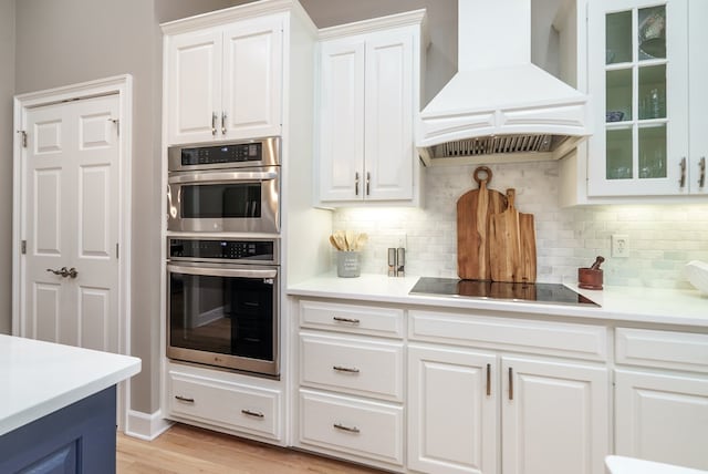 kitchen with white cabinetry, double oven, backsplash, black electric stovetop, and custom range hood