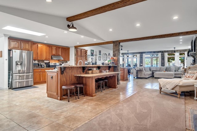 kitchen with a large island with sink, lofted ceiling with beams, appliances with stainless steel finishes, light tile patterned flooring, and a kitchen bar