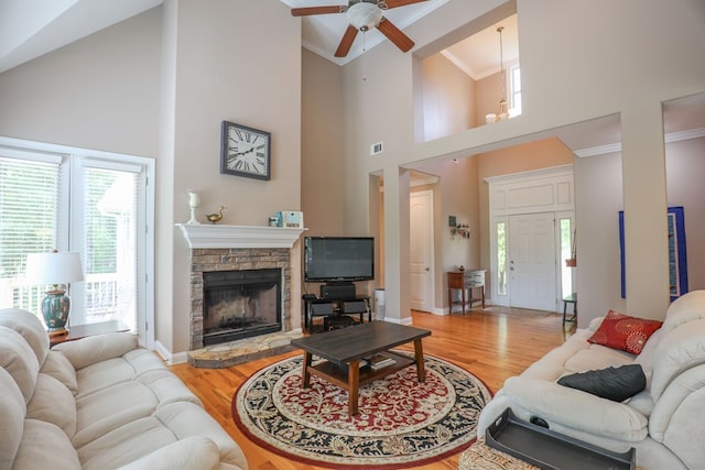 living room featuring ceiling fan, a high ceiling, a stone fireplace, light hardwood / wood-style flooring, and crown molding