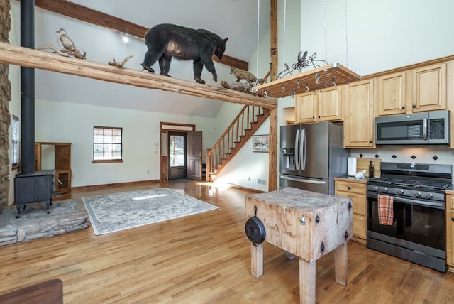 kitchen with a towering ceiling, stainless steel appliances, light brown cabinets, hardwood / wood-style floors, and a wood stove