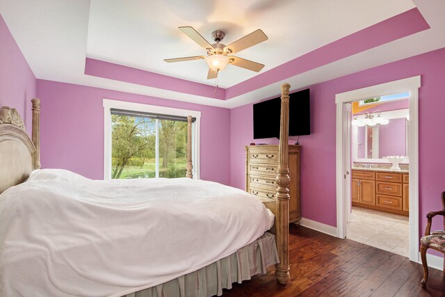 bedroom with a tray ceiling, dark wood-type flooring, connected bathroom, and ceiling fan