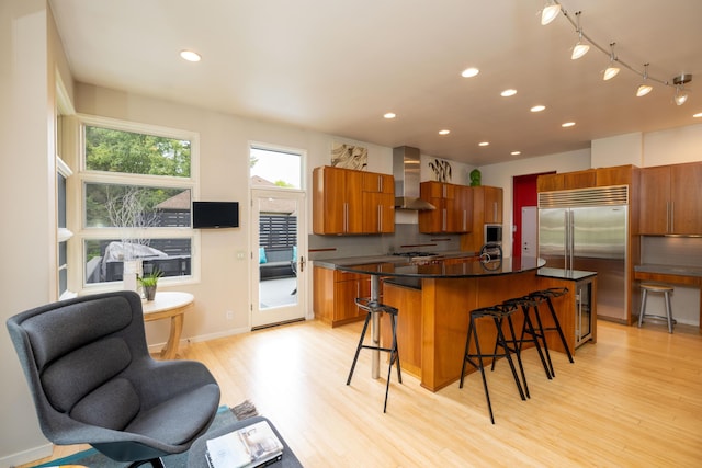 12737 Sanctuary Pl, Grand Haven, MI 49417 - kitchen with wall chimney exhaust hood, brown cabinetry, a breakfast bar, light wood-style flooring, and dark countertops
