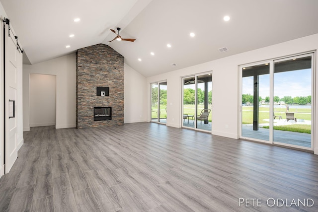 unfurnished living room featuring ceiling fan, hardwood / wood-style floors, high vaulted ceiling, a stone fireplace, and a barn door