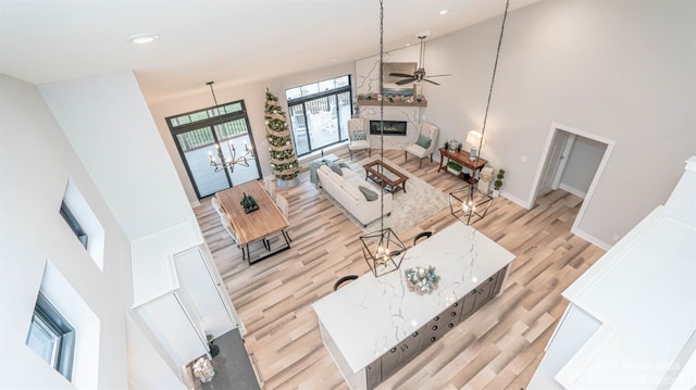 living room featuring a stone fireplace, light hardwood / wood-style flooring, ceiling fan with notable chandelier, and a high ceiling