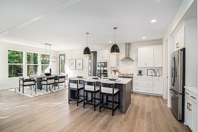 kitchen featuring appliances with stainless steel finishes, pendant lighting, an island with sink, white cabinets, and wall chimney exhaust hood