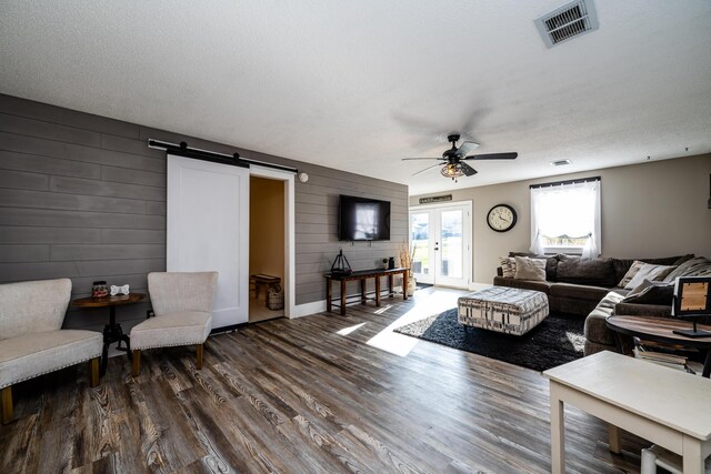 living room with a textured ceiling, dark hardwood / wood-style floors, a barn door, ceiling fan, and wooden walls