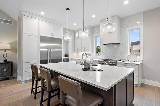 kitchen with stainless steel built in refrigerator, hanging light fixtures, an island with sink, custom range hood, and white cabinets