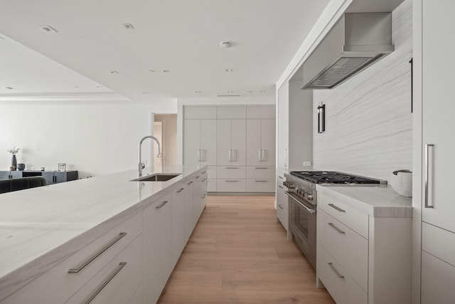 kitchen with light wood-type flooring, stainless steel stove, wall chimney exhaust hood, light stone countertops, and sink