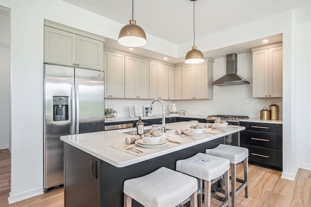 kitchen featuring wall chimney range hood, a kitchen island with sink, stainless steel appliances, light hardwood / wood-style floors, and decorative light fixtures