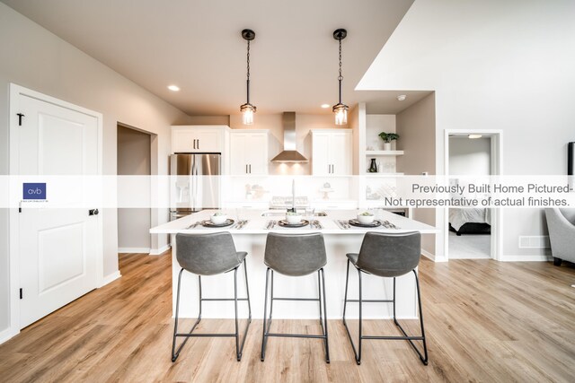 kitchen with stainless steel refrigerator, white cabinets, hanging light fixtures, and wall chimney range hood