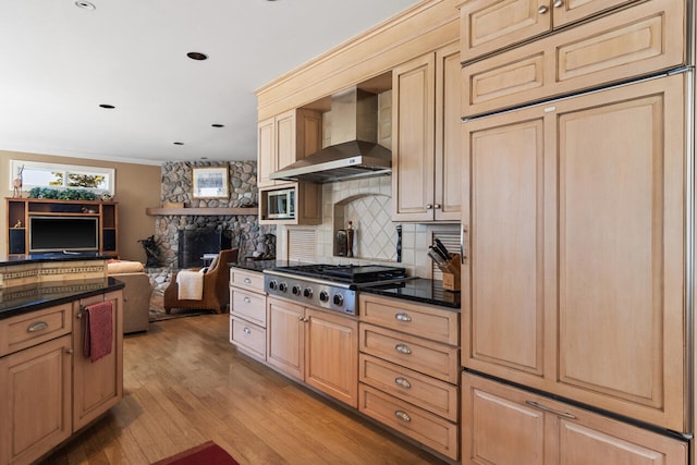 kitchen with wall chimney exhaust hood, a stone fireplace, tasteful backsplash, stainless steel gas stovetop, and light hardwood / wood-style floors