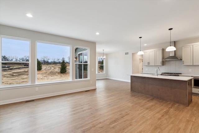 kitchen with decorative light fixtures, tasteful backsplash, light hardwood / wood-style floors, a kitchen island with sink, and wall chimney exhaust hood