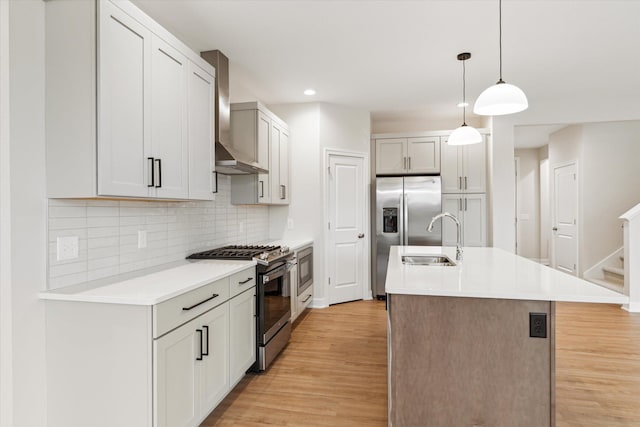 kitchen with sink, hanging light fixtures, an island with sink, stainless steel appliances, and wall chimney exhaust hood