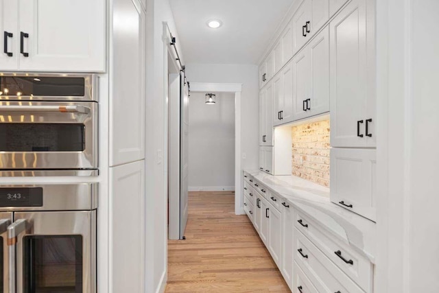 kitchen featuring double oven, white cabinets, light stone counters, a barn door, and light wood-type flooring