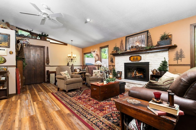 living room with lofted ceiling, ceiling fan with notable chandelier, a brick fireplace, hardwood / wood-style flooring, and a textured ceiling