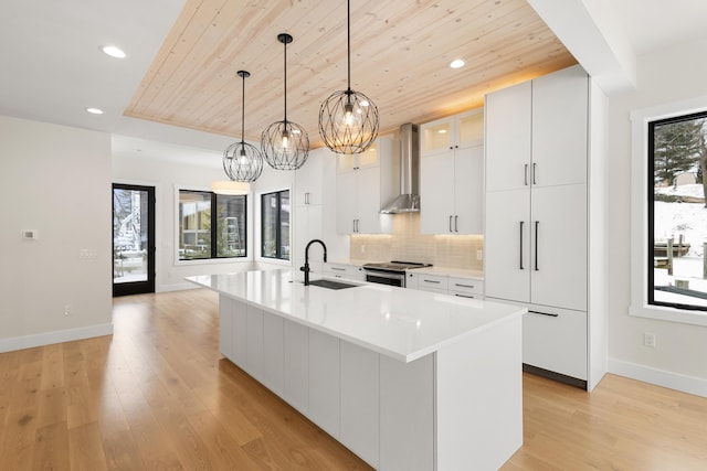 kitchen featuring wall chimney range hood, decorative light fixtures, sink, and a center island with sink