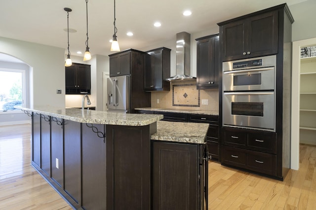 kitchen with an island with sink, hanging light fixtures, wall chimney range hood, and appliances with stainless steel finishes