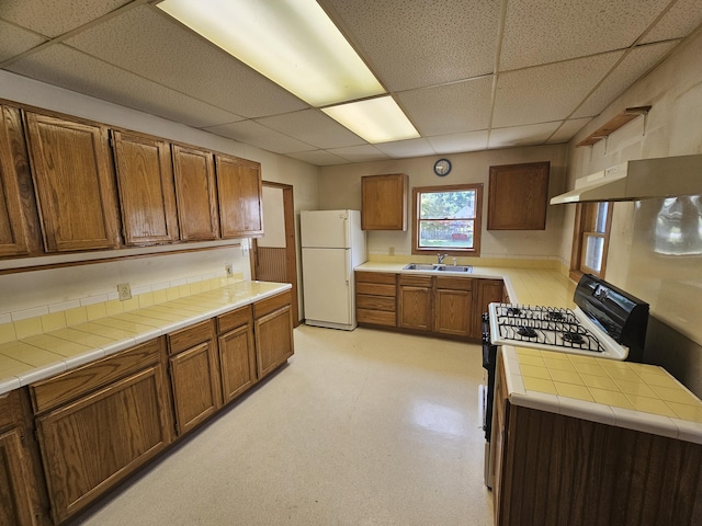 kitchen with a paneled ceiling, tile countertops, sink, white refrigerator, and gas range