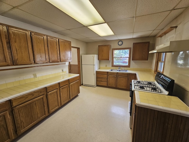 kitchen with a paneled ceiling, sink, white refrigerator, tile counters, and gas stove