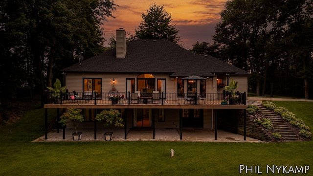 back house at dusk featuring a wooden deck, a patio area, and a lawn