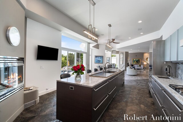 kitchen with light stone countertops, sink, dark brown cabinets, an island with sink, and ceiling fan