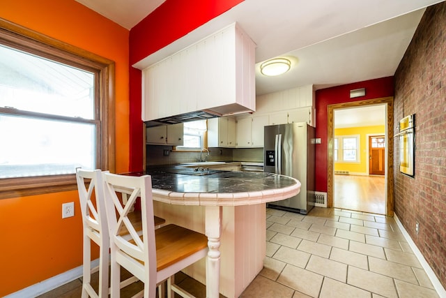 kitchen with tile countertops, white cabinets, stainless steel fridge, a kitchen bar, and brick wall