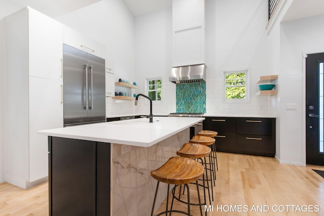 kitchen with light hardwood / wood-style flooring, a center island with sink, a healthy amount of sunlight, decorative backsplash, and wall chimney exhaust hood