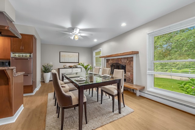 dining room with light hardwood / wood-style floors, a baseboard radiator, a stone fireplace, and ceiling fan