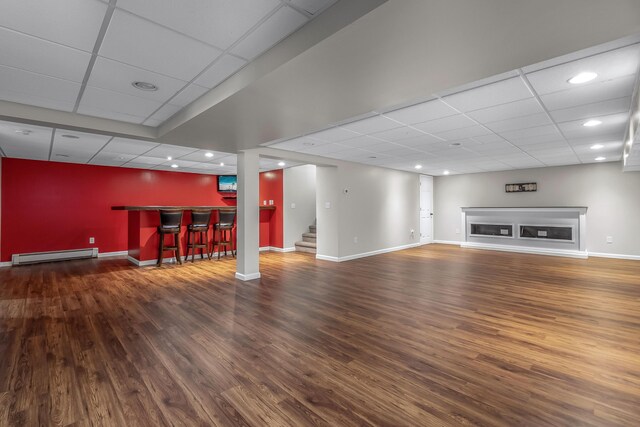 unfurnished living room featuring indoor bar, baseboard heating, a paneled ceiling, and dark wood-type flooring