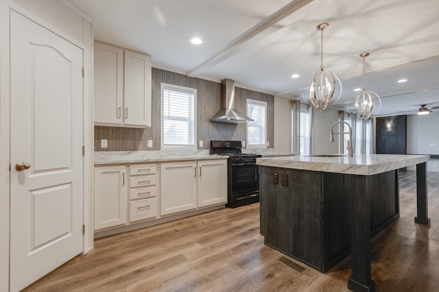 kitchen featuring ceiling fan with notable chandelier, an island with sink, light wood-type flooring, black / electric stove, and wall chimney exhaust hood