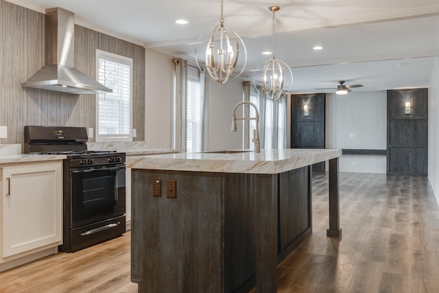 kitchen with ceiling fan with notable chandelier, black range, an island with sink, light hardwood / wood-style floors, and wall chimney range hood