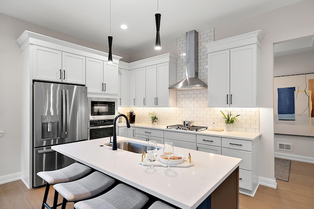 kitchen featuring a sink, wall chimney range hood, appliances with stainless steel finishes, light wood finished floors, and decorative backsplash