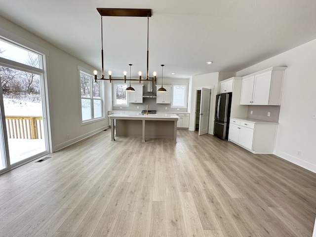 kitchen with white cabinetry, wall chimney exhaust hood, a kitchen island with sink, stainless steel refrigerator, and pendant lighting